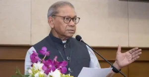 Older man with glasses speaking at a podium, holding a sheet of paper, with a purple orchid bouquet in front.