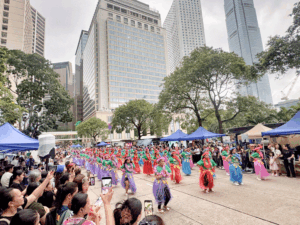 Crowd watches a colorful street dance with tai or hula-inspired performers in purple, red, and green skirts amid city skyscrapers and tents.