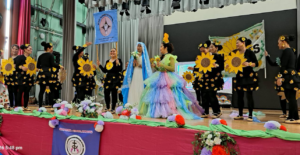 Group of performers on a stage, many in black outfits with sunflower decorations, surrounding a woman in a pastel rainbow tutu dress at center.
