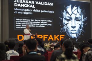 Speaker in white suit and pink belt presents on a stage with a large screen showing Indonesian text about digital risks; geometric face graphic on the right; seated audience in front.