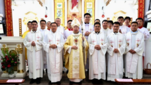Group of Catholic clergy in white robes posing with a bishop in gold vestments inside a church sanctuary, altar and ornate decorations in view.