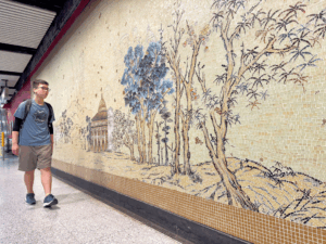 Young man with a backpack walks along a subway platform past a beige mosaic wall depicting trees and a pavilion.