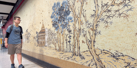 Young man with a backpack walks along a subway platform past a beige mosaic wall depicting trees and a pavilion.