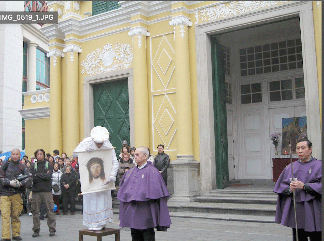Religious procession outside a yellow church: priests in purple robes with a man carrying a framed portrait of Jesus; crowd gathers nearby.
