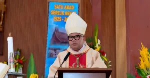 Religious leader in cream vestments and a tall mitre speaks at a lectern with a microphone, candle and flowers nearby in a church setting