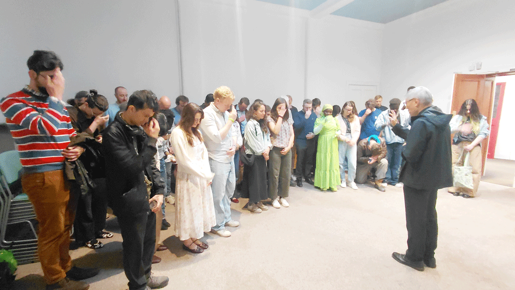 A diverse group of people standing in a line in a bright room, many with heads bowed as a man in a dark jacket speaks to them (audience moment).
