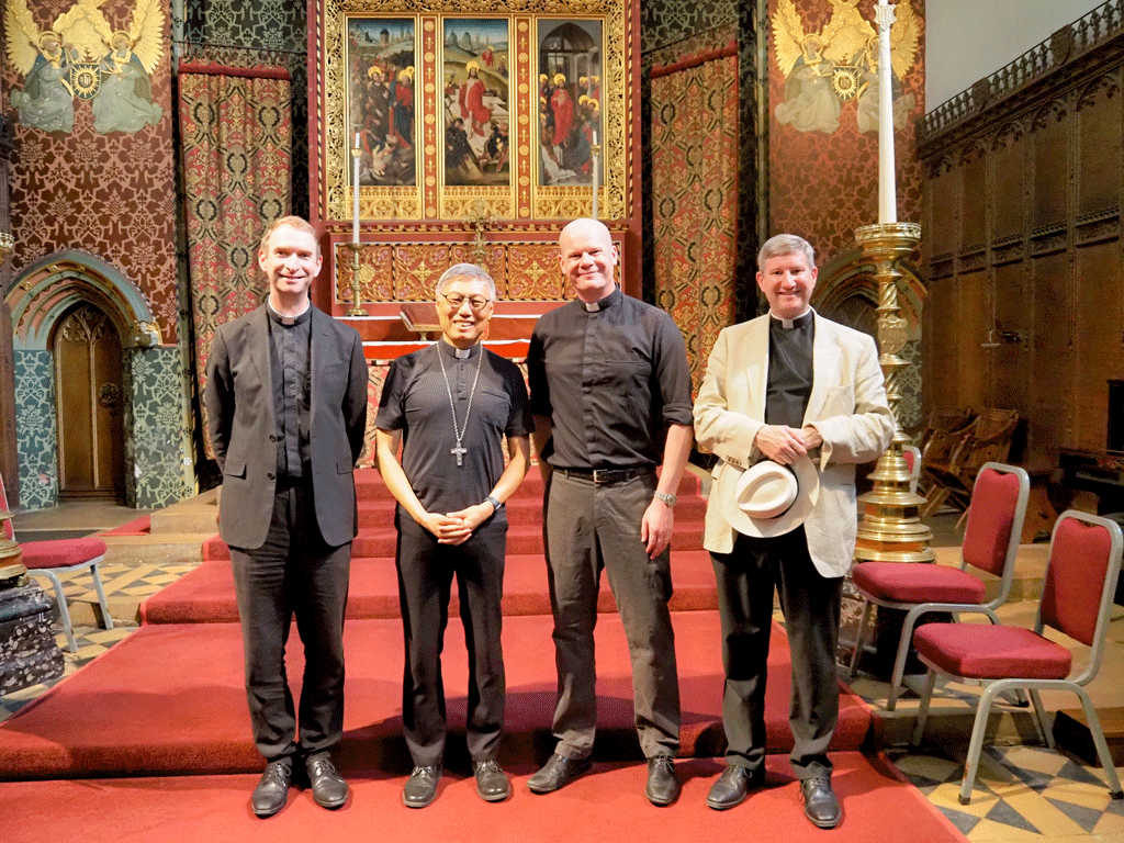 Four clergy members stand on a red-carpeted church altar area, smiling at the camera in ornate surroundings.