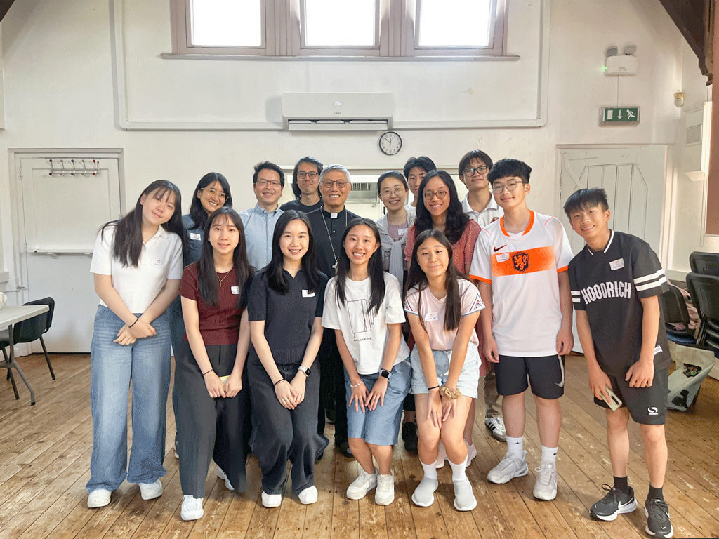 Group of diverse students and two adults posing for a photo in a sunlit room with wooden floor and clock on the wall.