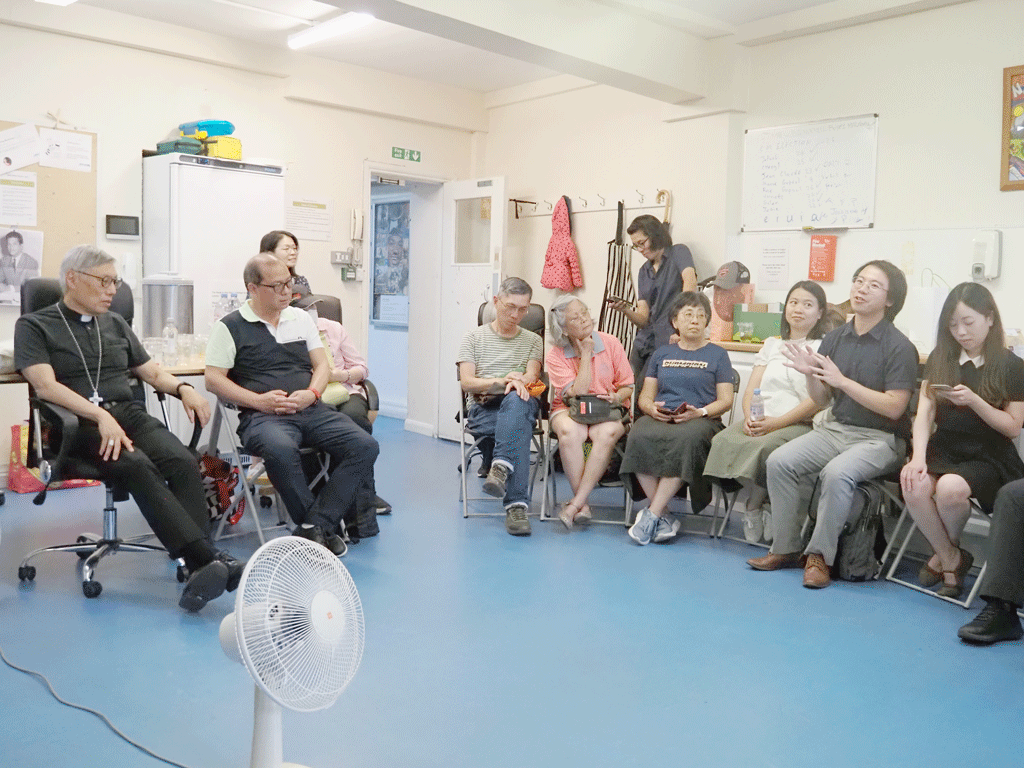 Group of adults seated in a circle in a bright room, a fan in the foreground, and people chatting softly.
