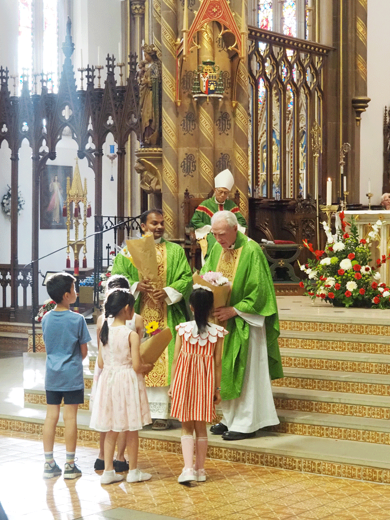 Priests in green vestments receive flower bouquets from four children during a liturgical ceremony in an ornate church interior.
