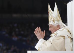 Pope in ornate white and gold vestments praying with hands together, facing left at a podium.