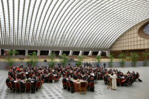 A large hall filled with cardinals in red robes seated at round tables for a formal banquet under a curved, striped glass ceiling with tall plants along the back wall.