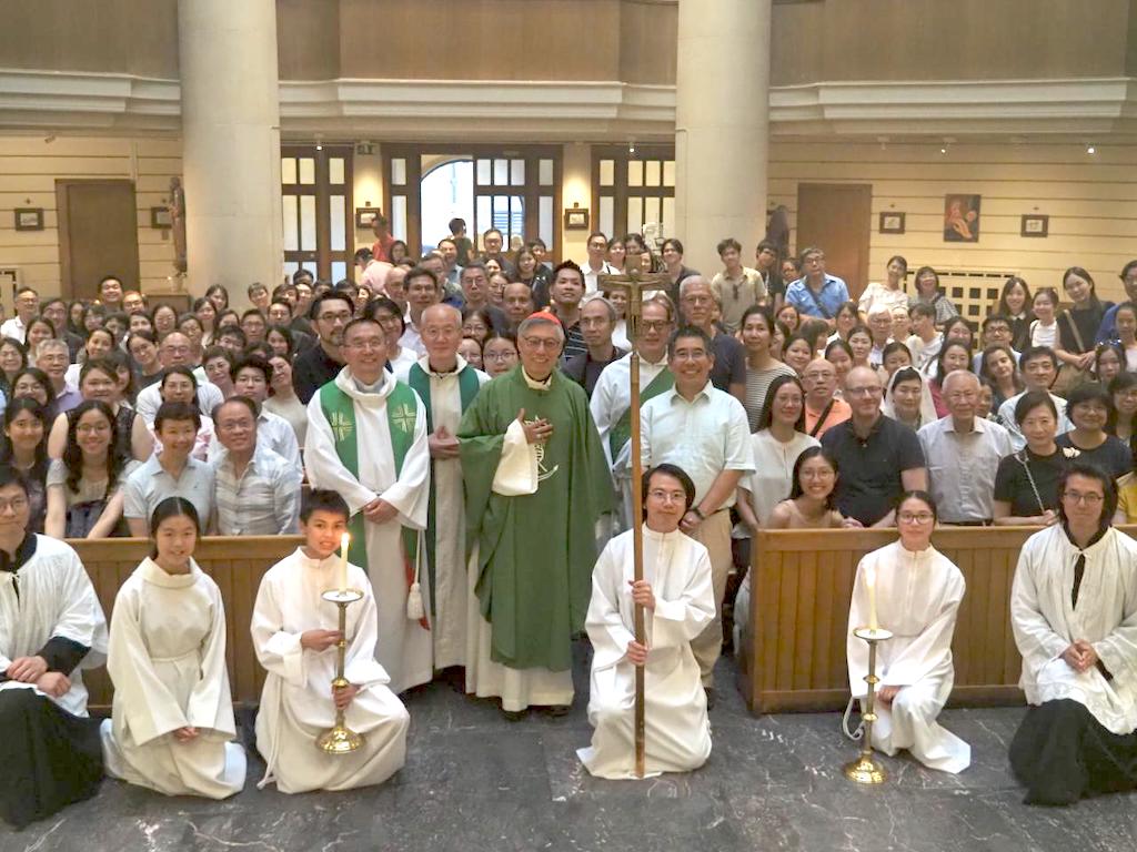 Large group of people in a church posing for a photo, with priests in green vestments and altar servers kneeling in front holding candles and a cross.