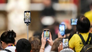 Crowd of people at an event holding up smartphones to record or take photos, some on selfie sticks, with yellow-shirted attendee in the foreground.