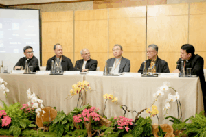 Six men in formal attire sit behind a long conference table on a panel, with microphones and nameplates in front and floral arrangements at the desk edge.