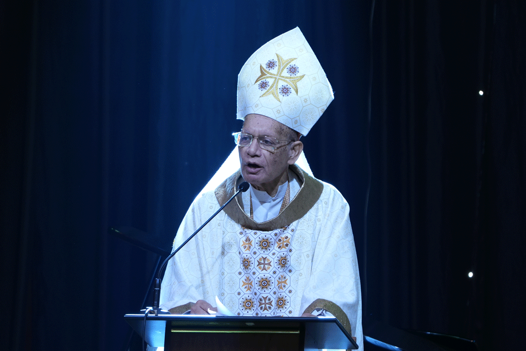 Clergy member in white liturgical vestments and mitre speaks at a pulpit with a mic, delivering a sermon.