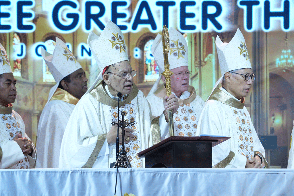 Group of bishops in white and gold vestments on a stage, one holds a ceremonial staff beside a lectern under a glowing backdrop.