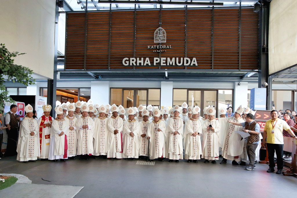 Group of Catholic clergy in white liturgical vestments and mitres posing for a photo outside a building labeled 'GRHA PEMUDA' with a cathedral emblem above.