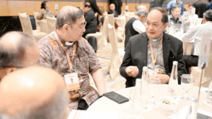 Two priests seated at a round banquet table, engaged in conversation during a formal event, with drinks and bowls on the table.