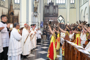 Clergy in white robes stand in a church as colorfully dressed dancers with feather headdresses perform in front of them.
