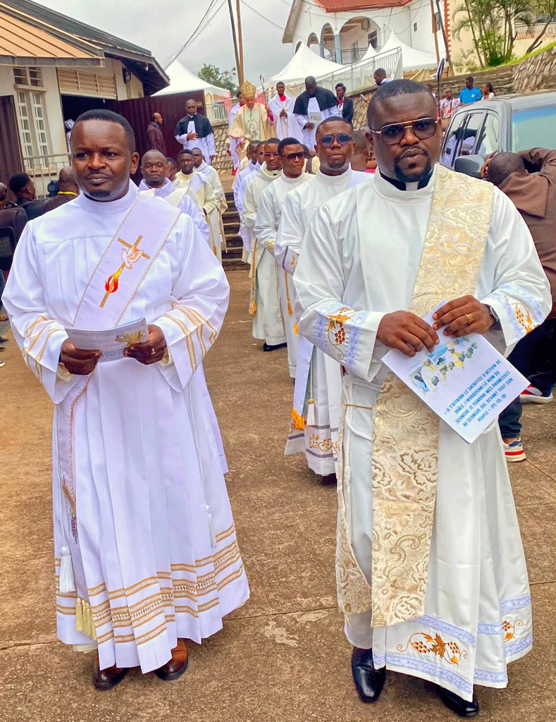 Line of priests in white liturgical robes with gold embroidery marching in a religious procession along a paved street, church and onlookers in the background.