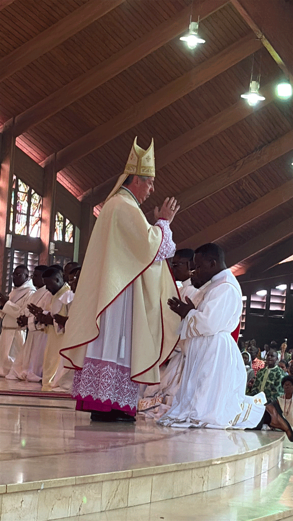 A bishop in cream vestments and a mitre blesses a kneeling man in white robes on a church stage, with helpers and attendees nearby.