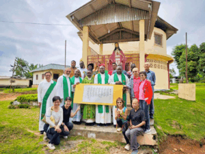 Group of clergy and volunteers pose with a framed plaque in front of a small church courtyard.