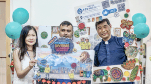 Three people pose with a large colorful poster board, surrounded by handmade crafts and teal balloons in a decorated room.