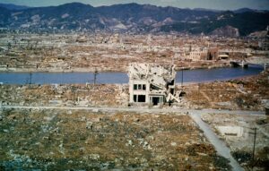 Wide view of a devastated city covered in rubble, a river in the foreground, and a few ruined buildings with mountains behind them—evocative of post‑war destruction.