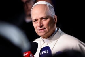 Close-up of a senior religious leader in white robes speaking to reporters, with multiple microphones in front and a solemn, grimacing expression.