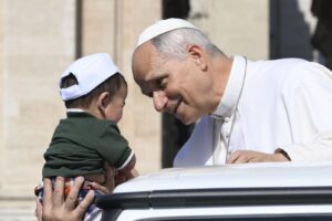 Pope in white robes leaning to greet a toddler held by an adult outdoors beside a white car.