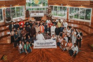 Group photo of diverse men, women, and children in a wood-floored hall with an altar and large windows; a banner at the front reads 'Global Chaplains, Animators Formation and Exchange (CAFÉ) Program 2026'.