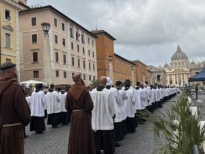 Line of people in white robes with brown-robed figures along a cobblestone street toward St. Peter's Basilica, Vatican City.
