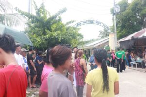 Crowd of people gathered outside the Jose National High School entrance arch, with market stalls and media volunteers nearby.