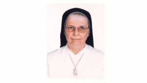 Portrait of a Catholic nun wearing a white habit, black veil, glasses, and a pendant necklace, head-and-shoulders portrait.