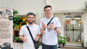 Two men smiling and giving thumbs up outside a building next to a vertical banner and glass door.