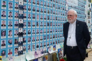 Elderly clergyman with white hair and glasses stands before a blue wall covered with dozens of portrait cards with names and photos.