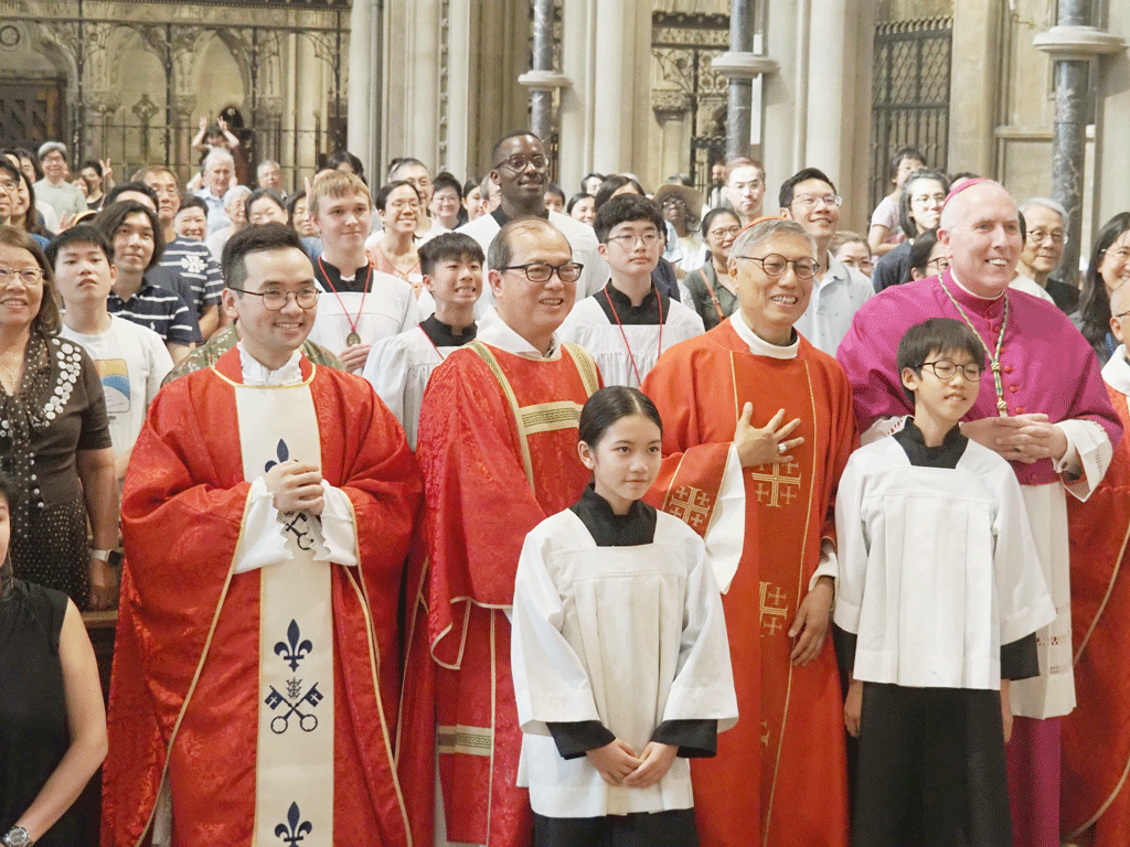 Catholic priests in red vestments pose with two altar servers in white albs inside a church, smiling at the camera, with a crowd behind them.