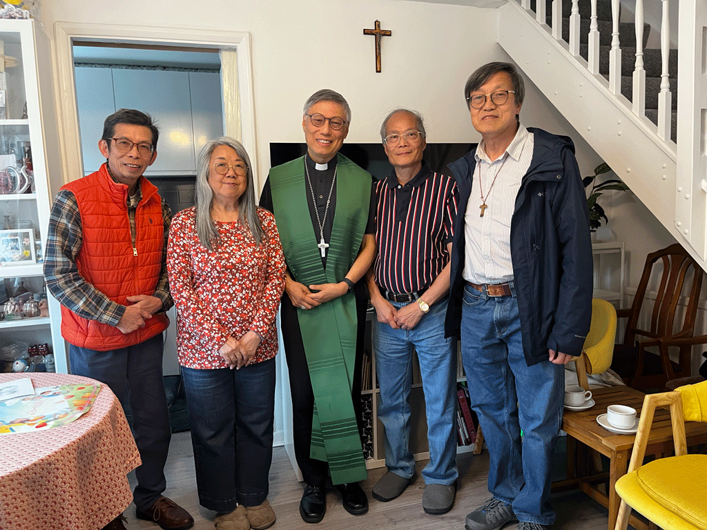 Five adults posing in a living room; a priest in a green stole stands center, flanked by two men and two women, with a cross on the wall behind them.