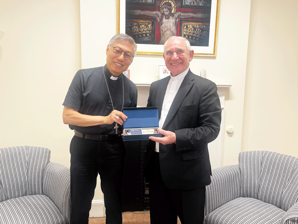 Two men, one in clerical collar and one in a suit, smile while presenting a blue box containing items in a living-room setting.