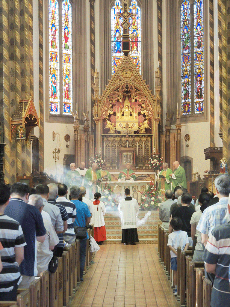 Mass in a church with priests in green vestments at the altar, and a congregation standing in pews.