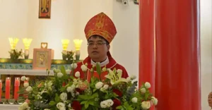 Priest in red ceremonial vestments and a tall mitre stands behind a large flower arrangement at a church altar with lit candles.