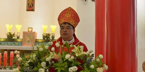 Priest in red ceremonial vestments and a tall mitre stands behind a large flower arrangement at a church altar with lit candles.
