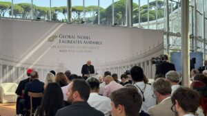 Speaker at a podium in front of a banner reading Global Nobel Laureates Assembly on Artificial Intelligence and Nuclear War, with an attentive audience and cameras nearby.