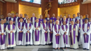 Group of Catholic priests in white robes with purple stoles posing for a group photo inside a church.