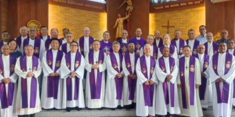 Group of Catholic priests in white robes with purple stoles posing for a group photo inside a church.
