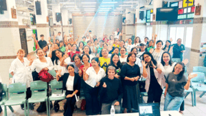 Large diverse group posing for a group photo in a bright indoor hall, many waving and smiling at the camera.