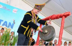 Man in colorful traditional dress strikes a large gong with a mallet at a cultural ceremony inside a tent.
