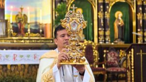 Priest in white and gold vestments holding a large ornate gold monstrance during a Catholic ceremony inside a church.