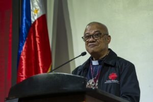 Clergyman with glasses at a podium, addressing an audience; Philippines flag behind him.